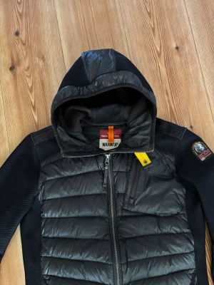 Svart Parajumpers cardigan - Snygg svart cardigan från Parajumpers med huva och dragkedja framtill. Jackan har quiltad front, släta ärmar och en gul detalj vid halsen. Märkespatch på vänster ärm och bröstficka med dragkedja. Perfekt för våren. Nypris 4500