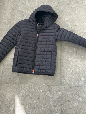 Svart pufferjacka med huva - Snygg svart pufferjacka med huva och dragkedja framtill. Jackan har två sidofickor och quiltad design som ger en modern look. Perfekt för kalla dagar och enkel att matcha med olika outfits. Orange detaljer på dragkedjan för en cool touch.