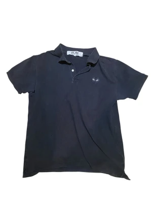 Svart piké från Comme des Garçons - Svart pikétröja från Comme des Garçons Play med klassisk krage, korta ärmar och två knappar framtill. På bröstet sitter det ikoniska hjärtemärket med ögon. Tillverkad i bomull för en skön och avslappnad känsla. Storlek L men passar M också