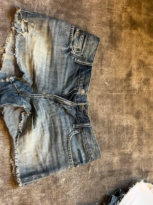 Blå jeansshorts från Polo Ralph Lauren - Snygga blå jeansshorts från Polo Ralph Lauren med råa kanter och slitna detaljer. Klassisk femficksmodell med knappgylf och låg midja. Perfekta för sommaren och ger en avslappnad vibe till din outfit. Storlek 14, motsvarar storlek 34/36