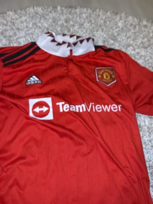 Manchester United Ronaldo matchtröja - Säljer en röd Manchester United matchtröja från Adidas med TeamViewer-logga på bröstet och Ronaldo 7 tryckt på ryggen. Tröjan har vita och svarta detaljer, klubbmärke på bröstet och klassisk krage med mönster. Perfekt för fotbollsfans!
