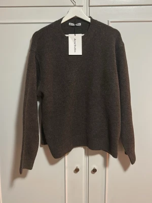 Brun stickad tröja från Acne Studios - Mörkbrun stickad tröja från Acne Studios med rund halsringning och ribbade muddar vid ärmslut och nederkant. Tröjan har en enkel, klassisk design och är tillverkad i ett mjukt material som passar perfekt för kyligare dagar.