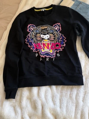 Svart sweatshirt med tiger från Kenzo - Svart sweatshirt från Kenzo med ikoniskt broderat tigerhuvud i färgglada nyanser och texten 'Kenzo Paris' på bröstet. Tröjan har rund hals, ribbade muddar och är tillverkad i mjukt bomullsmaterial.