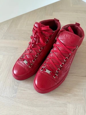 Balenciaga herr skor  - Snygga röda high-top sneakers i äkta läder med grov struktur. Skorna har metallöglor och matchande röda skosnören samt detaljer i silver framtill. Perfekta för dig som vill sticka ut med en färgstark och trendig look.