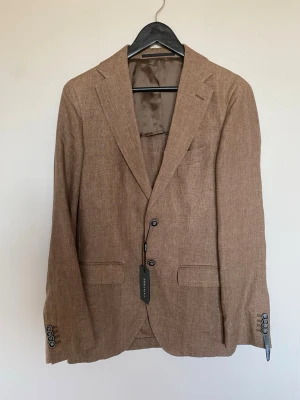 Brun linnékavaj från Cavaliere  - Snygg brun kavaj från Cavaliere i lätt linnematerial. Klassisk passform med två knappar framtill, dubbla fickor och dekorativa knappar vid ärmsluten. Perfekt för en stilren och avslappnad look.