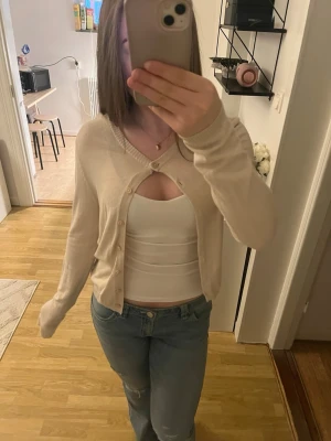 Beige cropped kofta med knappar - Snygg beige cropped kofta med v-ringning och knäppning framtill. Koftan har långa ärmar och en mjuk, tunn stickning som passar perfekt till jeans eller kjol. Enkel och stilren design utan synligt märke.