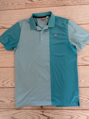 Tvåfärgad pikétröja från Puma - Snygg pikétröja från Puma i två nyanser av blått. Tröjan har klassisk krage, knäppning med tre knappar och Puma-logga på bröstet. Tillverkad i ett lätt och sportigt material som andas. Storlek S, mått 43 axel till axel och 73 cm lång