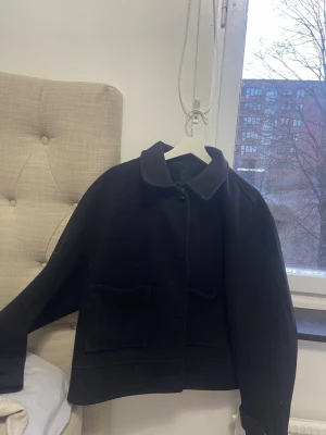 Svart ullkappa med fickor - Stilren svart kappa i ullmaterial med klassisk krage och två stora framfickor. Kappan har rak passform och knäppning framtill. Perfekt för dig som gillar minimalistisk och tidlös stil.