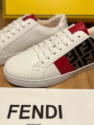 Vita Fendi sneakers med röda detaljer - Snygga vita sneakers från Fendi med röda detaljer på plösen och klassiskt Fendi-mönster på hälen. Skorna har en låg profil, vita snören och är tillverkade i slätt läder. Perfekta för dig som vill ha en clean och exklusiv look.