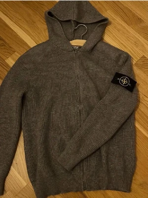 Grå stickad hoodie från Stone Island - Säljer en grå stickad hoodie från Stone Island med dragkedja framtill och ikonisk svart patch med logga på vänster ärm. Tröjan har ribbstickad struktur, huva och långa ärmar. Perfekt för dig som gillar streetwear och snygga detaljer.