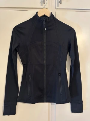 Svart träningsjacka från Lululemon - Snygg svart träningsjacka från Lululemon med figurnära passform, hög krage och hel dragkedja framtill. Jackan har två fickor med dragkedja och tumhål vid ärmsluten. Tillverkad i ett stretchigt funktionsmaterial som passar perfekt till träning.