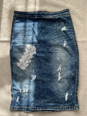 Blå jeanskjol med spetsdetalj - Säljer en blå denimkjol med slitningar och en snygg vit spetsapplikation på framsidan. Kjolen har en rak passform och är i klassisk jeansmodell med coola detaljer som ger en trendig look.