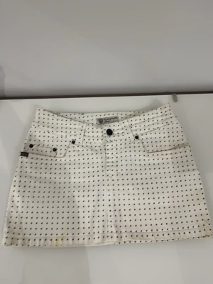 Vit prickig kortkjol från tiger if Sweden jeans  - Säljer en vit kortkjol med svarta prickar och klassisk jeanslook. Kjolen har fem fickor, bälteshällor och knappstängning framtill. Perfekt för dig som gillar en lekfull och trendig stil.