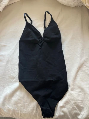 Svart body med smala axelband - Snygg svart body med smala, justerbara axelband och lätt rynkning vid bysten. Tillverkad i mjukt stretchigt material för skön passform. Perfekt att styla under skjortor eller tröjor för en clean look.