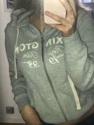 Grå hoodie från Lexington - Snygg grå zip-hoodie från Lexington med vit text och tryck framtill. Hoodien har dragsko i huvan, två fickor och ribbade muddar vid ärmar och nederkant. Perfekt för en avslappnad och sportig look.