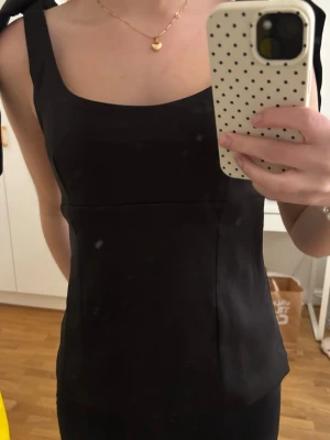 Svart linnetopp med rosetter - Snygg svart linnetopp med breda axelband som knyts upptill. Toppen har en rak, något figurnära passform och fyrkantig ringning. Perfekt att styla med jeans eller kjol för en trendig look.