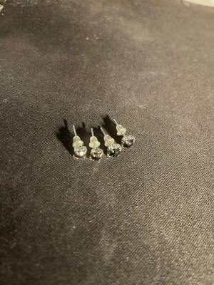 Silvriga örhängen med svarta stenar - Säljer två par små silvriga örhängen med gnistrande klara stenar. Klassisk rund form och stilren design som passar till det mesta. Örhängena har metallstift och transparenta bakstycken. Perfekta för dig som gillar enkla men snygga smycken.  OBS! Materialet är okänt, kan ej garantera att det är rostfritt.