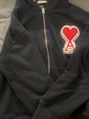 Svart hoodie från Ami - Säljer en svart hoodie från Ami Paris med dragkedja och stor röd hjärtlogga med bokstaven A på bröstet. Hoodien har huva med snörning och känguruficka framtill. Tillverkad i mjukt bomullsmaterial som känns skönt mot huden.