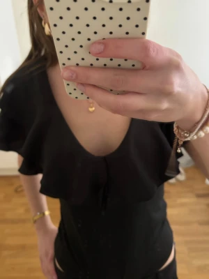 Svart topp med volang och öppen rygg - Snygg svart topp med djup v-ringning, volangdetaljer och öppen rygg. Modellen är figurnära och har korta ärmar med volang. Perfekt för dig som vill ha en trendig och stilren look. Stor i storleken så S/M