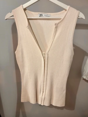 Beige ribbad ärmlös topp från Zara - Säljer en beige ribbad ärmlös topp från Zara med v-ringning och dragkedja framtill. Perfekt till jeans eller kjol för en enkel men snygg look. Materialet är mjukt och stretchigt för skön passform.