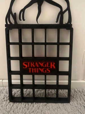 Stranger Things väggställ svart - Säljer ett svart väggställ med Stranger Things-logga i rött. Stället har rutmönster och en siluett av en Demogorgon på toppen. Perfekt för att visa upp samlarprylar eller dekorationer för fans av serien.