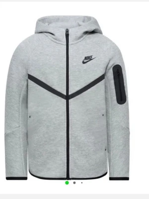 Grå Nike Tech Fleece hoodie - Snygg grå hoodie från Nike med dragkedja framtill och svart Nike-logga på bröstet. Hoodien har svarta detaljer, ficka på ärmen och en modern, sportig design. Tillverkad i mjukt fleece-material och har huva för extra komfort.Den är helt nytt är använde bara en gång priset kan diskuteras.