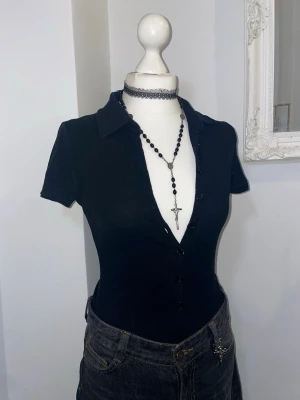 Svart ribbad kortärmad bodysuit - Snygg svart ribbad bodysuit med korta ärmar och krage. Modellen är figurnära med knappar framtill och så djup v-ringning som man vill. Köpt i London. 