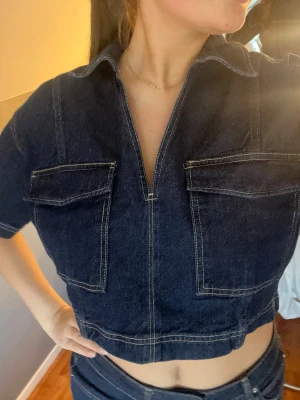 Mörkblå croppad jeansskjorta - Trendig croppad skjorta i mörkblå denim med korta ärmar och två stora bröstfickor. Skjortan har en djup v-ringning och markerade sömmar i kontrastfärg. Perfekt till ett par höga jeans för en cool och avslappnad look.