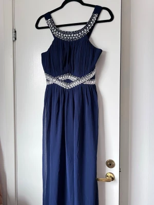 Marinblå balklänning med pärlor och stenar, storlek M - En marinblå balklänning med vackra pärldetaljer runt halsringningen och midjan. Plisserad design och breda axelband ✨ dubbelt tyg och går hela vägen ner till fötterna. Endast använd en gång! 💕 Midja: ca 70-75cm - Längd: 140cm