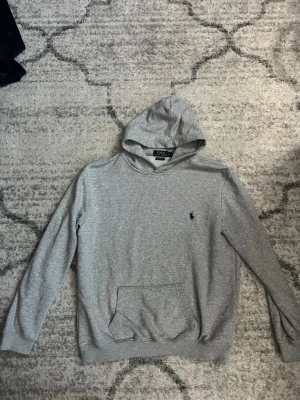 Grå hoodie från Polo Ralph Lauren - Snygg grå hoodie från Polo Ralph Lauren med klassisk marinblå logga broderad på bröstet. Tillverkad i mjuk bomull, har huva och en stor magficka. Perfekt för en chill och stilren look.