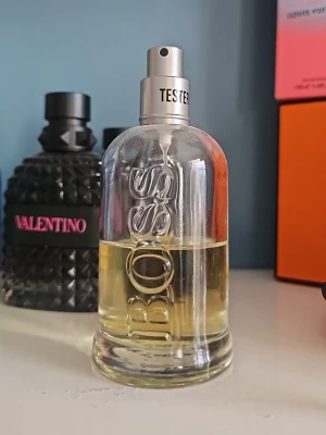 Hugo Boss parfymflaska - Klassisk parfym från Hugo Boss i en genomskinlig glasflaska med silverfärgad spraykork märkt 'TESTER'. Flaskan har en stilren design med BOSS-loggan i relief på framsidan och innehåller en ljusgul vätska.