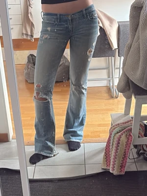 Ljusblå slitna bootcut jeans - Säljer ett par ljusblå bootcut jeans med flera slitningar och hål på benen för en trendig, avslappnad look. Jeansen har klassisk femficksmodell, låg midja och är tillverkade i denim. Perfekta för dig som gillar en edgy stil.