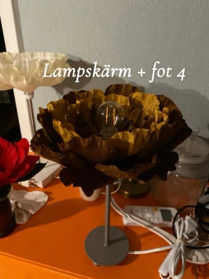 Lampa i form av en blomma  - Lampskärm + fot ingår) egentillverkad.  Höjd: ca 35 cm Bredd: ca 26 cm Viktigt att veta:  1. Lampan tål inte direkt solljus, då kommer färgen med störst sannolikhet att blekas tyvärr. Beror dock lite på hur stark solen är där ni placerar den. Men den är ej passande för t ex fönsterbrädan. Den passar bäst vid ett nattduksbord, mörkare rum där solen ej når fram direkt, eller t ex en byrå mitt i ett rum.  2. Glödlampa kommer ej att medfölja. Jag rekommenderar att köpa en av de svagast lamporna. 