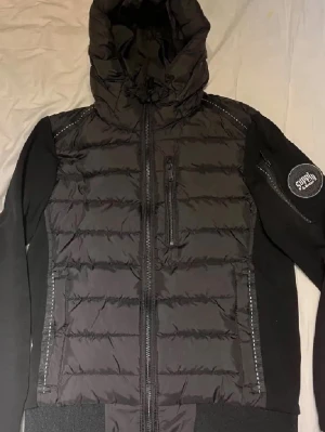 Svart pufferjacka med huva Supply - Snygg svart pufferjacka från Supply med huva och dragkedja framtill. Jackan har vadderad kropp, mjuka ärmar och detaljerade sömmar. Bröstficka med dragkedja och logga på ärmen ger en cool streetwear-look. Perfekt för kalla dagar.