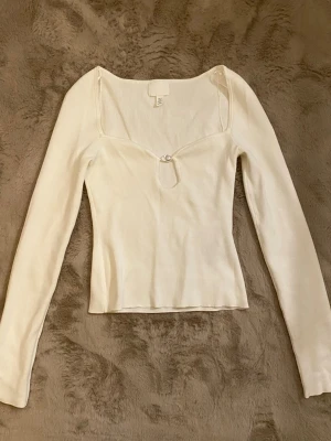 Vit långärmad topp med pärldetalj - Vit långärmad topp med smickrande v-ringning och en söt pärldetalj framtill. Toppen har en figurnära passform och är tillverkad i ett mjukt, stretchigt material som känns bekvämt mot huden. Perfekt att styla med jeans eller kjol.