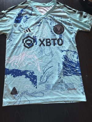 Inter Miami Messi t-shirt blå Adidas - Snygg ljusblå Inter Miami t-shirt från Adidas med Messi 10 på ryggen. Tröjan har korta ärmar, v-ringning och coola blå och rosa grafiska mönster. Tillverkad i polyester och perfekt för fotboll eller streetwear.