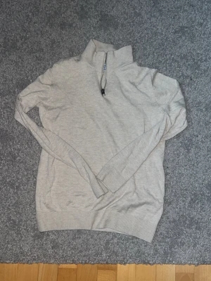 Beige stickad tröja med halv dragkedja - Snygg beige stickad tröja med hög krage och halv dragkedja framtill. Tröjan har långa ärmar och ribbade muddar vid ärmslut och nederkant. Perfekt för lager på lager och enkel att matcha med olika outfits.