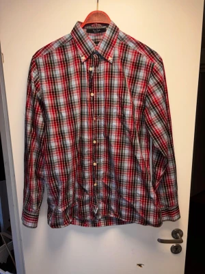 Rutig röd skjorta från gant - Säljer en klassisk långärmad skjorta från Gant i rött, vitt och svart rutigt mönster. Skjortan har knappar framtill, bröstficka och button down-krage. Perfekt för dig som gillar en tidlös och avslappnad stil.