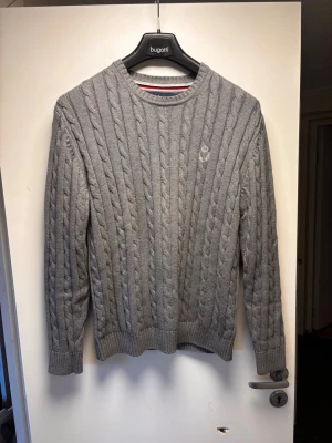 Grå kabelstickad tröja carl william - Snygg grå kabelstickad tröja med rund halsringning och diskret logga på bröstet. Tröjan har långa ärmar och ribbade muddar vid ärmslut och nederkant. Perfekt för dig som gillar klassisk och stilren design.