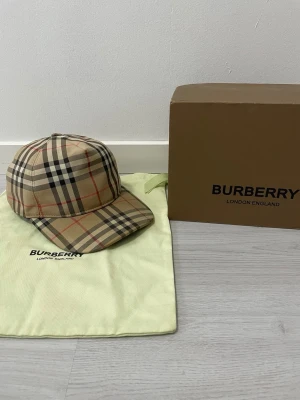 Burberry keps - Säljer en klassisk Burberry keps i beige med det ikoniska rutmönstret i svart, vitt och rött. Kepsen har böjd skärm och justerbar rem bak. Tillverkad i bomull och levereras med originalpåse och box. St M använd 
