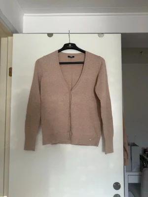 Beige kofta med v-ringning - Säljer en stilren beige kofta med v-ringning och långa ärmar. Koftan har en enkel design med knäppning framtill och ribbade muddar vid ärmslut och nederkant. Perfekt att slänga över en t-shirt eller topp för en clean look.
