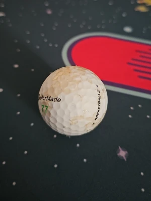 Golfboll TaylorMade Rocketballz - En vit golfboll från TaylorMade med texten 'ROCKETBALLZ' och siffran 77 i grönt. Bollen har klassisk dimple-yta och är tillverkad i slitstarkt material, perfekt för dig som gillar golf och vill ha en boll med bra känsla.