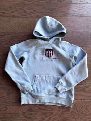 Ljusblå hoodie från GANT - Snygg ljusblå hoodie från GANT med broderad logga på bröstet. Hoodien har en klassisk känguruficka och ribbade muddar vid ärmar och nederkant. Perfekt för en avslappnad och sportig stil. Finns en liten fläck på en armbågen. 