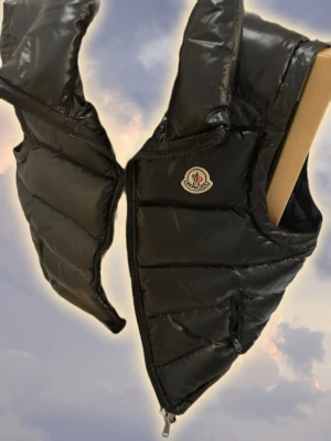 Svart exklusiv puffer-dunväst  - Snygg svart dunväst med glansig finish och klassisk logga på bröstet. Västen har hög krage, två dragkedjeförsedda fickor och quiltad design. Perfekt för lager-på-lager och streetstyle.