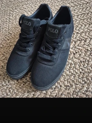 Svarta Polo Ralph Lauren sneakers - Stilrena svarta sneakers från Polo Ralph Lauren med canvasöverdel och detaljer i svart läder. Klassisk låg modell med snörning och logga på sidan samt plös. Perfekta för en clean och avslappnad look.