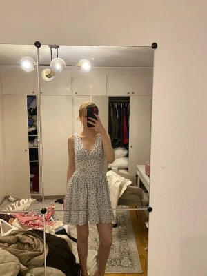 Vit blommig kortklänning v-ringad - Söt vit kortklänning med blått blommönster och v-ringning. Klänningen är ärmlös och har en lätt utställd kjol som ger en luftig känsla. Perfekt för varma dagar och har en feminin, ungdomlig stil.