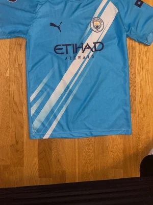 Manchester City matchtröja Puma - Snygg ljusblå Manchester City matchtröja från Puma med vita diagonala ränder, klubbmärke och Etihad Airways-logga på bröstet. Nummer 7 och namnet Marmoush på ryggen. Perfekt för fotbollsfans som vill sticka ut på planen eller läktaren.