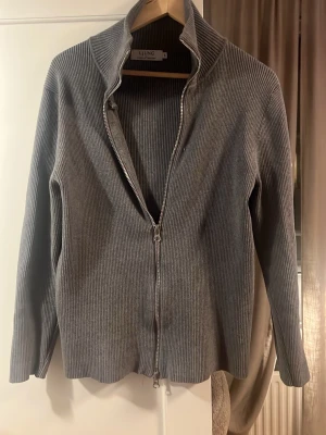 Grå ribbad ziptröja från Llung - Snygg grå ribbad tröja från Llung med dragkedja hela vägen och hög krage. Tröjan har lång ärm och är tillverkad i ett mjukt, stretchigt material som ger en clean och modern look. Perfekt för lager på lager och enkel att styla.