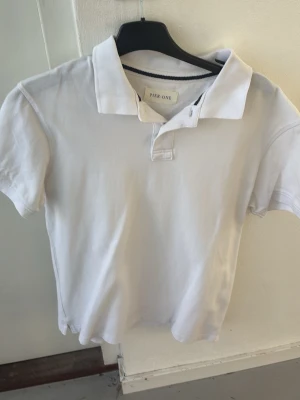 Vit pikétröja från Pier One - Klassisk vit pikétröja från Pier One med krage och korta ärmar. Tröjan har en enkel knappslå vid halsen och är tillverkad i mjuk bomull som känns skön mot huden. Perfekt till jeans eller shorts för en clean och stilren look. Storlek S men passar även M
