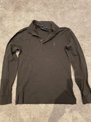 Ralph lauren långärmad piké - Klassisk svart långärmad skjorta från Ralph Lauren med lila broderad logga på bröstet. Skjortan har krage, knappar vid halsen och är tillverkad i mjuk bomull. Perfekt för en stilren och avslappnad look.